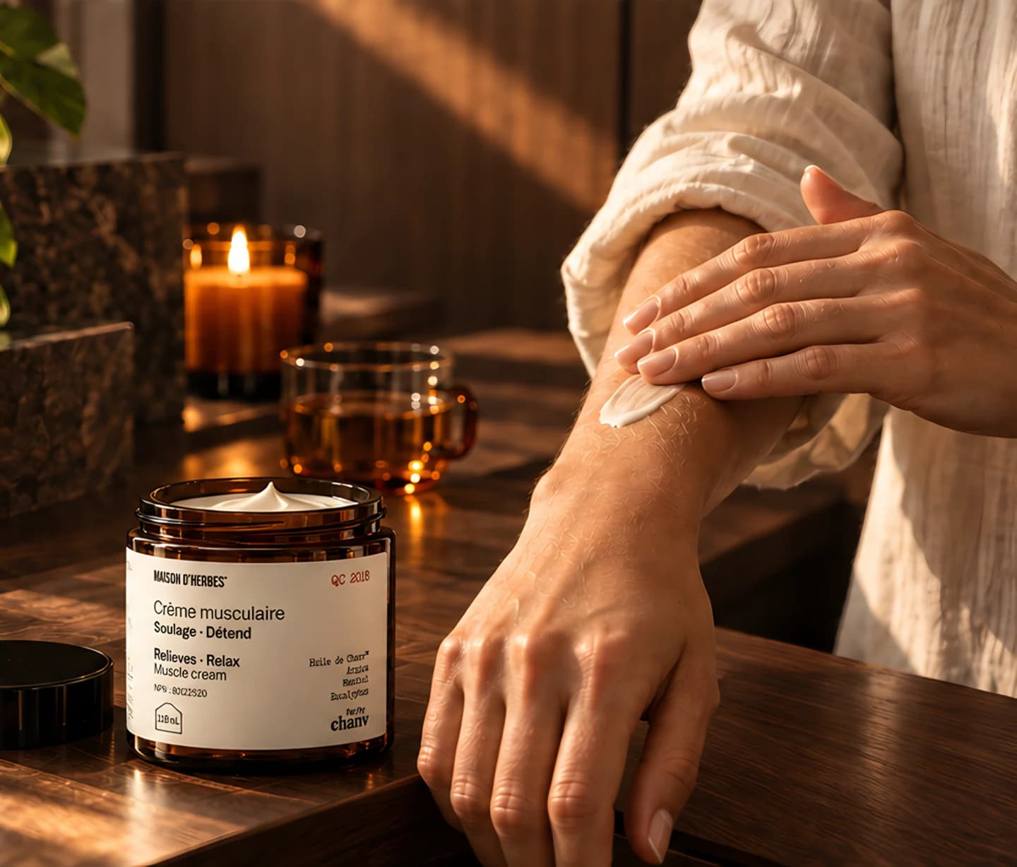 Une main applique la Crème musculaire Maison d’Herbes sur un avant-bras, près d’une bougie allumée.