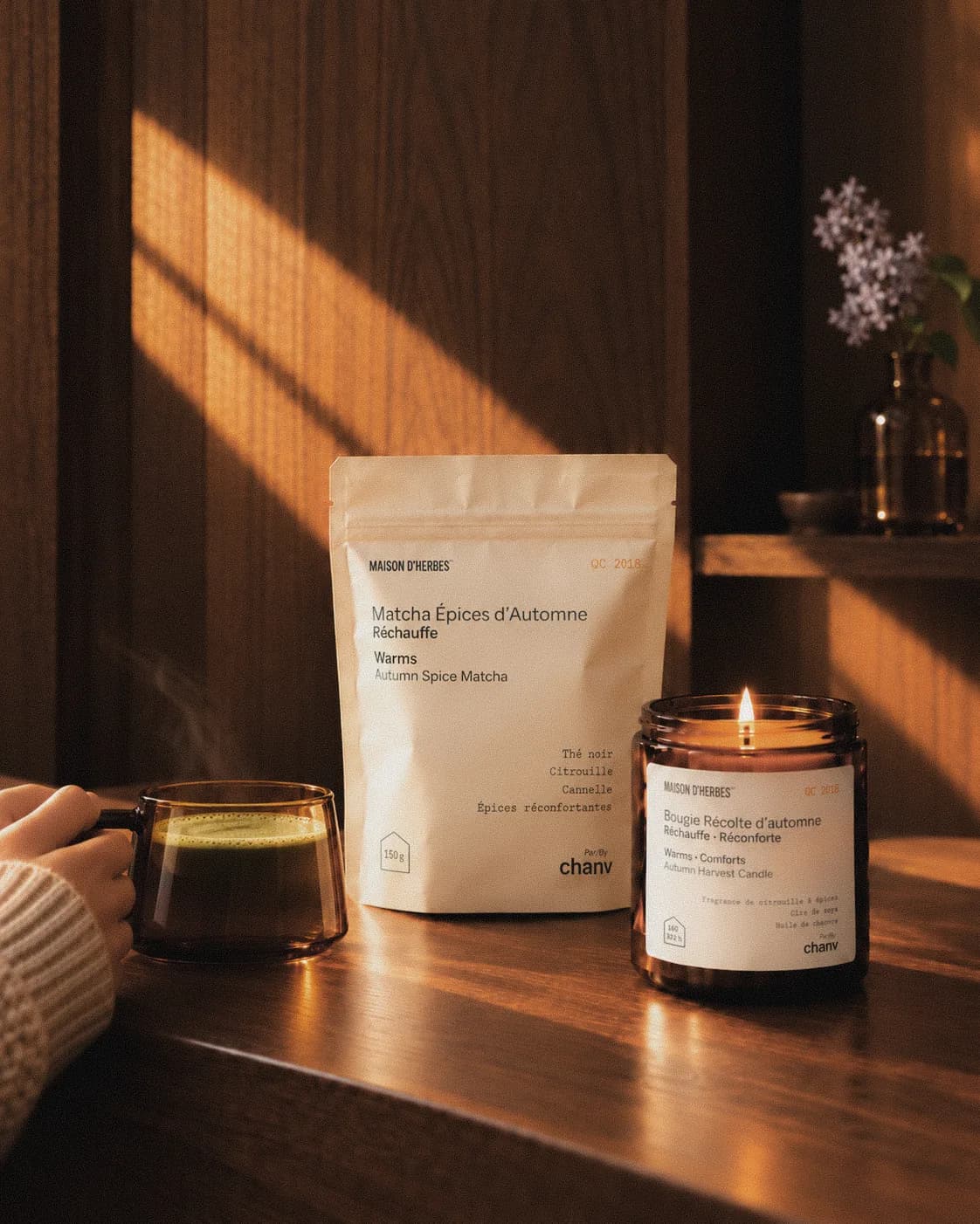 Une main tient une tasse de thé devant un sachet de Matcha Épices d’Automne et une bougie Récolte d’automne Maison d’Herbes allumée, sur une table de bois dans une lumière chaude