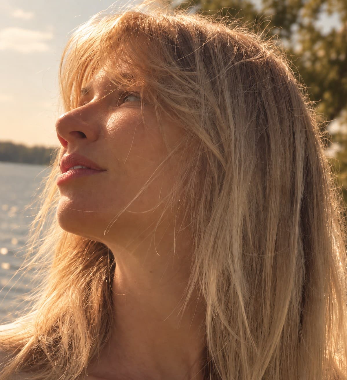 Visage levé vers la lumière dorée du couchant, cheveux blonds au vent devant un plan d’eau