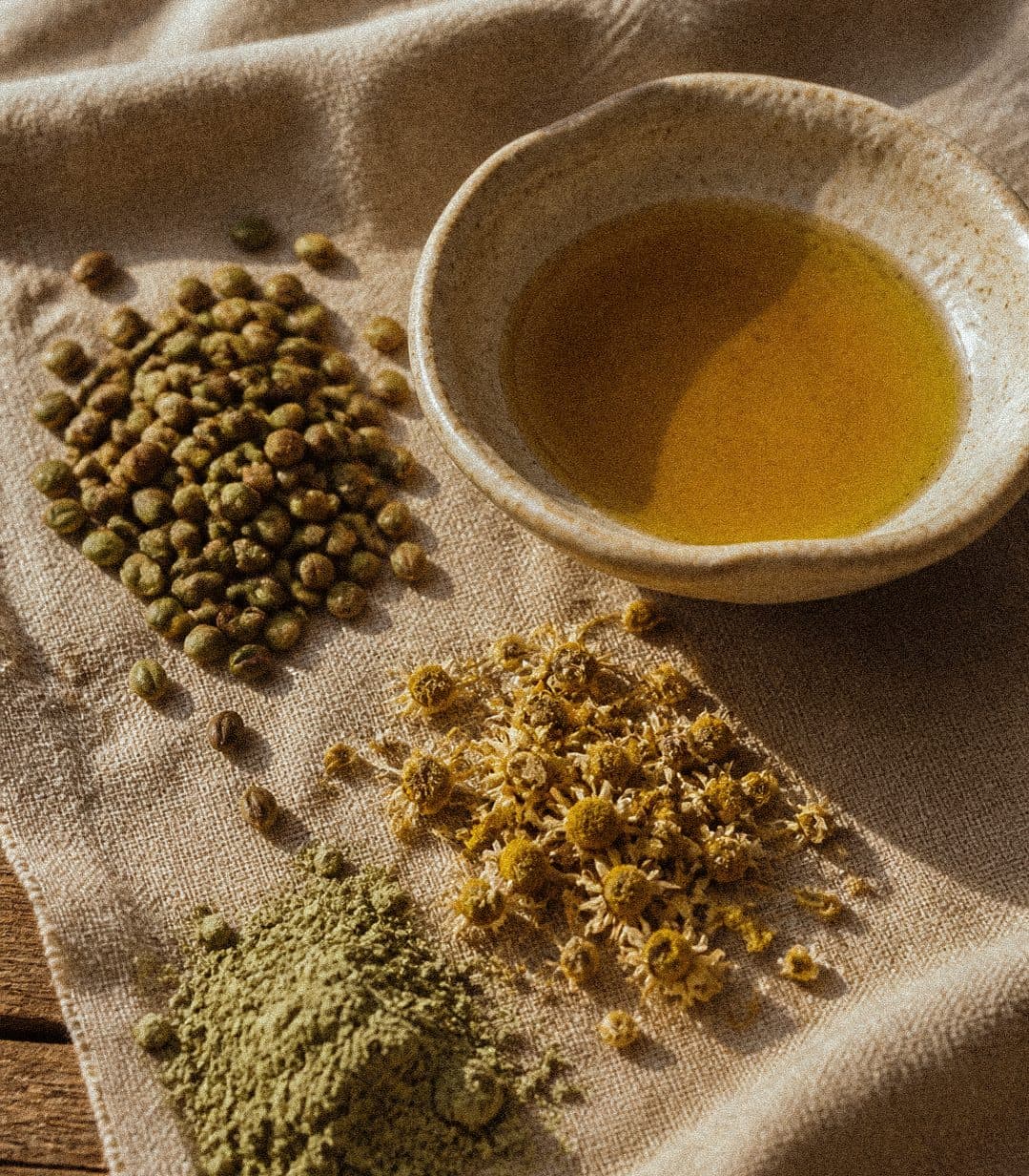 Sur un lin froissé, un tas de graines de chanvre, des fleurs de camomille séchées, une poudre de plante verte et un bol de grès rempli d’huile dorée