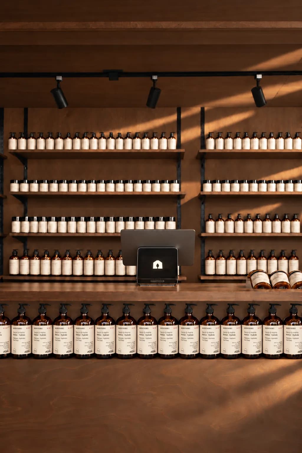 Comptoir de bois de la boutique baigné de lumière, bordé de savons à mains en flacons ambrés, devant un mur de rayonnages garnis